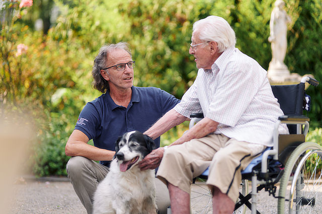 alt="Pflegekraft kommuniziert mit einem Senior, beide streicheln dabei einen Hund"