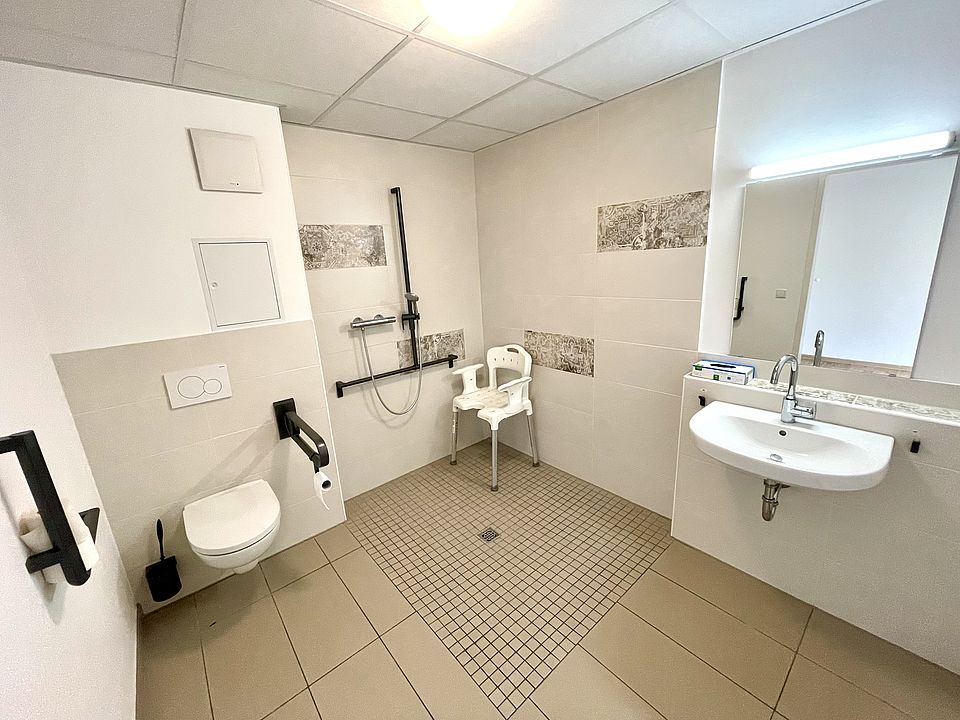 alt="Barrierefreies Badezimmer mit ebenerdiger Dusche, Haltegriffen und unterfahrbarem Waschbecken"