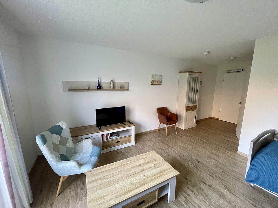 alt="Helles, möbliertes Zimmer mit Bett, Fernseher und Sessel"